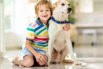 Egy kisfiú és egy kutya együtt ülnek a lakásban, boldogan mosolyognak.