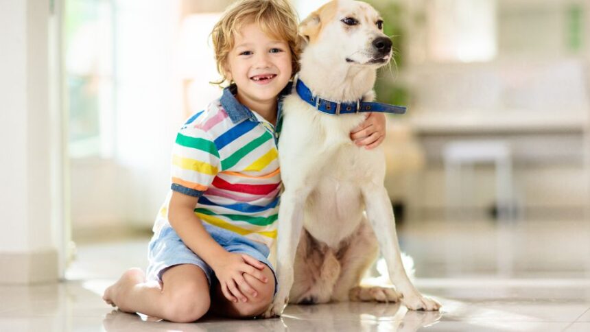 Egy kisfiú és egy kutya együtt ülnek a lakásban, boldogan mosolyognak.