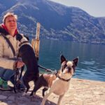 Egy férfi kutyákkal áll a vízparton, gyönyörű hegyekkel a háttérben.