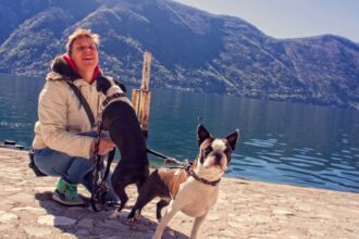 Egy férfi kutyákkal áll a vízparton, gyönyörű hegyekkel a háttérben.