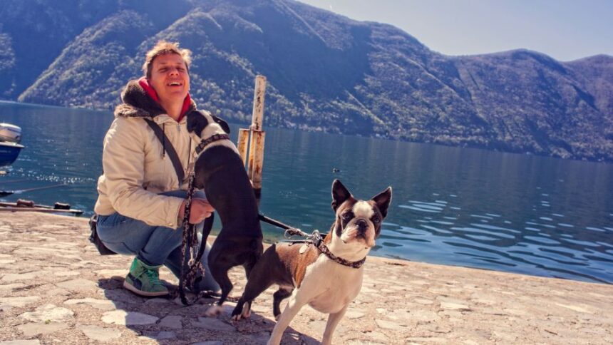 Egy férfi kutyákkal áll a vízparton, gyönyörű hegyekkel a háttérben.