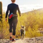 Egy nő kutyájával sétál a naplementében, gyönyörű természetben.