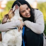 Egy nő boldogan öleli kutyáját, miközben a szabadban ülnek.